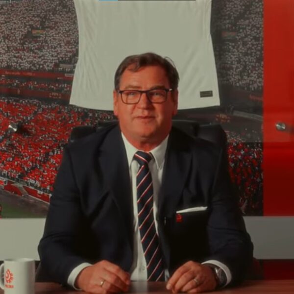 Jan Urban w garniturze siedzi przy biurku na tle dużego zdjęcia stadionu piłkarskiego wypełnionego kibicami w biało-czerwonych barwach