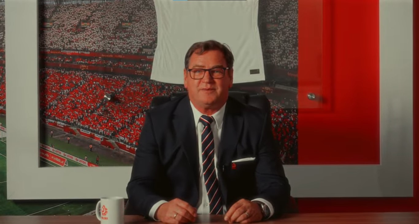 Jan Urban w garniturze siedzi przy biurku na tle dużego zdjęcia stadionu piłkarskiego wypełnionego kibicami w biało-czerwonych barwach