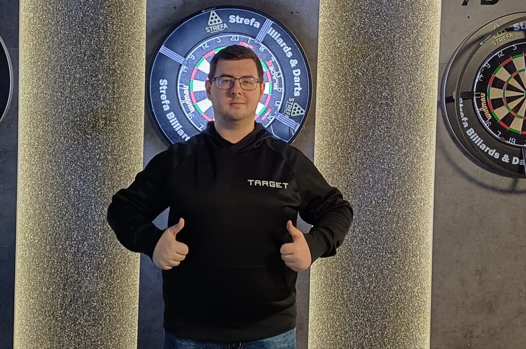 Sebastian Białecki stojący na tle tarczy z kciukami w górę